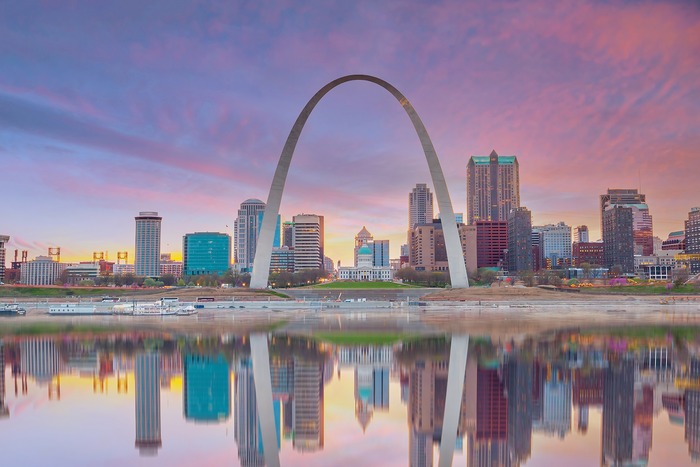 The Gateway Arch reflected in the Mississippi River at sunset, with the St Louis skyline behind it.